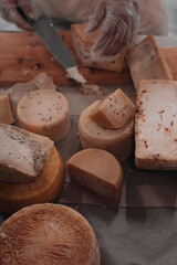 Man's hand with knife cutting a cheese on the wooden board on the table in the kitchen. Healthy eating and lifestyle. Various types of cheese. Toned