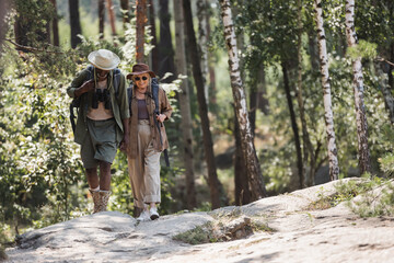 Smiling elderly woman with backpack walking near african american husband in forest.