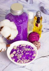 Fototapeta na wymiar Lavender-scented sea salt crystals in a bottle, white bowl on a white table, face body care oil bottles, cosmetic essential serum, cotton flower. Spa beauty day. Beautiful composition. Vertical photo.