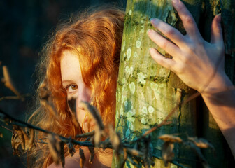 Portrait of beautiful sexy young woman, redhead hugging tree trunk in forest with friendly smile in...