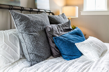 Closeup of decorative pillows cushions in shams and white bedding comforter duvet on bed in bedroom with illuminated lamp on nightstand and bright natural light from window