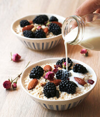Healthy oat meal cereal as breakfast with blackberries fruit and almonds with whole grains, dried pink rose. While hand holding bottle of milk pouring into a bowl, clean food concept  