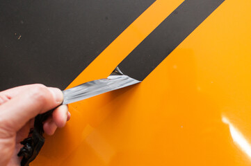 A man removes sticky stripes of old black foil from the hood of an orange car.
