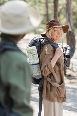 Cheerful elderly hiker with backpack and binoculars looking at blurred african american husband.