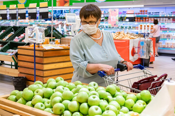 An elderly woman in a medical mask chooses apples in the store. Grandma in the fruit section of the supermarket. .