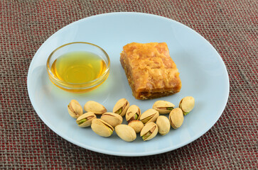 Piece of baklava with honey in glass bowl and pistachio nuts in shells on blue snack plate