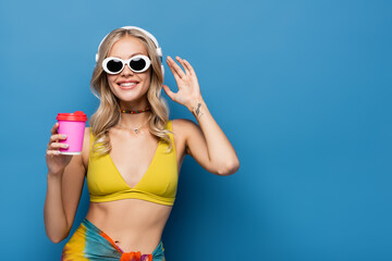 smiling young woman in wireless headphones and bikini top holding paper cup on blue.