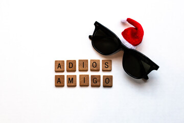 Adios Amigo wood block letters. A pair of folded black wayfarer-style sunglasses with a small Santa red-white Christmas hat on white. Holiday-themed, Christmas in July stock image. Vacation. Centred.