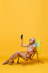 smiling woman in sunglasses and swimsuit sitting in deck chair with cocktail and smartphone on...