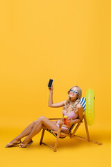 smiling woman in sunglasses and swimsuit sitting in deck chair with cocktail and smartphone on yellow.