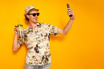 male tourist traveler in hat, Hawaiian shirt, sunglasses on vacation with drink in cocktail glass takes selfie on phone, communicates via video link. isolated on yellow background with space for text