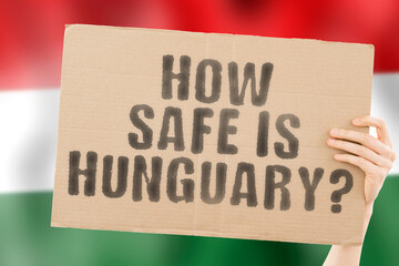 The question " How safe is Hungary? " on a banner in men's hand with blurred Hungarian flag on the background. Safety. Street. Outdoor. Dangerous. Security. Attack. Criminal. Criminality