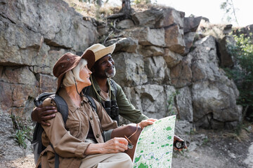 Cheerful african american hiker with compass hugging smiling wife with map near rock.