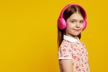 Adorable kid girl in stylish summer clothes, listening to music in headphones and looking at camera on yellow background