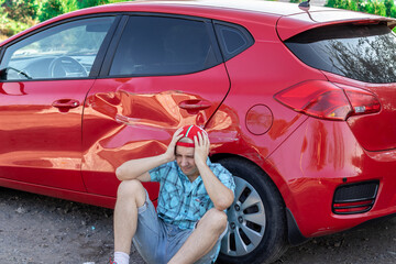a young man sits upset near his car with a dent after an accident - obrazy, fototapety, plakaty