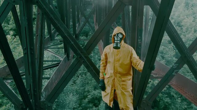 A man in a protective suit, gas mask and black leather gloves stands alone against the backdrop of a green forest under the bridge. The atmosphere of the post-apocalypse, after a man-made disaster.