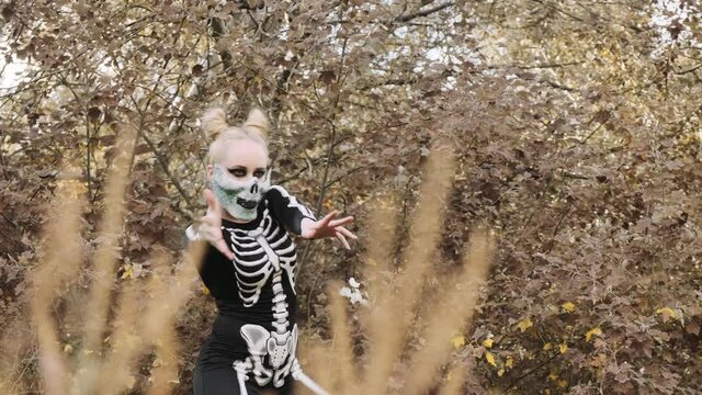 A girl with a scary make-up in a skeleton costume, in a mask with rhinestones in a gloomy autumn forest dances and moves scary. Close-up. Foreshortening through the grass. Halloween