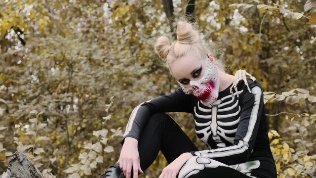 A girl with a scary make-up in a skeleton costume, a mask with rhinestones in a gloomy autumn forest. Sits on a withered tree. The skeleton's hand falls on the shoulder. Halloween