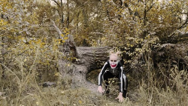 A girl with a scary make-up in a skeleton costume, a mask with rhinestones in a gloomy autumn forest. Jumps and crawls scary to the camera. Halloween, autumn holiday concept. Slow motion, 4K footage