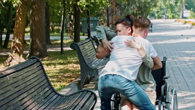 A guy helping disabled woman to sit on a bench from her wheelchair