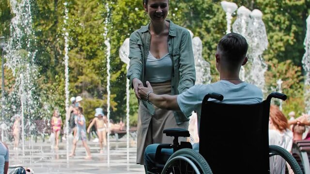 Sad man in a wheelchair sitting in front of a park and his happy girlfriend trying to cheer him up