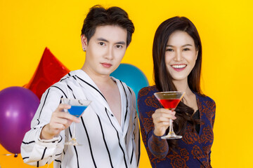 Portrait studio shot of Asian male and female teenager lover couple model in fashionable wears standing posing smiling in party look at camera holding cocktail tall glasses drink on yellow background