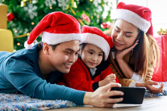 Happy Asian family father mother and daughter wears sweater with red and white Santa Claus hat lay down on carpet floor using smartphone camera taking selfie photo celebrating Christmas eve together