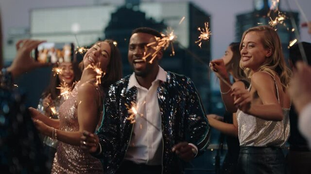 happy fashion friends dancing with sparklers celebrating new years eve at glamorous rooftop party friend using smartphone sharing video of celebration on social media 4k