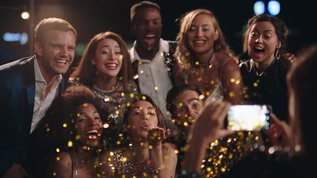 happy group of friends posing for photo celebrating reunion party wearing stylish fashion enjoying glamorous evening together blowing confetti at smartphone camera sharing on social media  