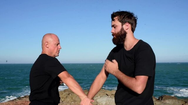 two men in black practice kung fu punches and kicks. wrestling sports training on the beach by the sea. Rocky coast, rugged landscape. men are fighting