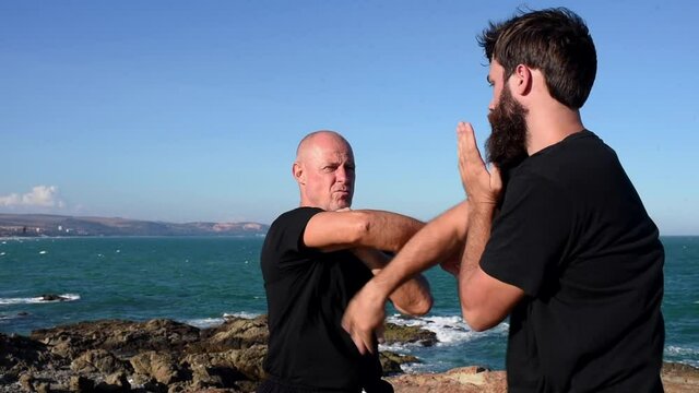 two men in black practice kung fu punches and kicks. wrestling sports training on the beach by the sea. Rocky coast, rugged landscape. men are fighting. malibu 2 man fight