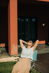Portrait of calm woman sunbathing in backyard. Mid adult lady wearing sunglasses sitting on chair at her house. Resting outdoors concept