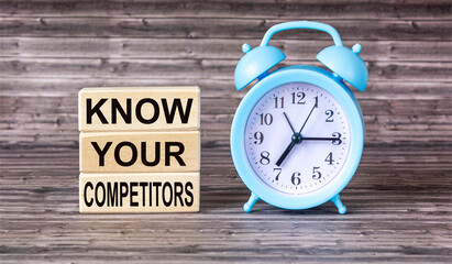 The text KNOW YOUR COMPETITORS is written on wooden blocks and on a wooden background with an alarm clock beside