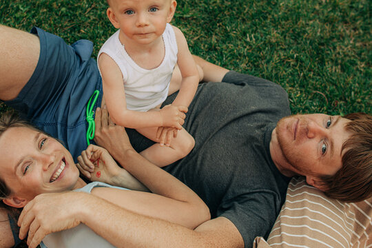 Portrait Of Happy Family With Little Daughter Lying On Grass. Couple With Toddler Resting On Front Yard. Happy Family Concept