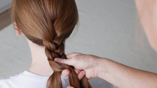 Close up view of making braid on red hair girl. Mother braids for her daughter. Process of making hairstyle for daughter. Process of braiding of red hair.