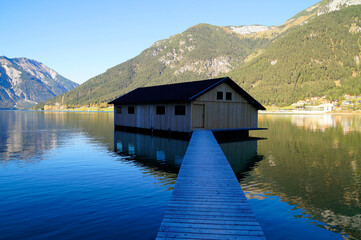 a wooden pier leading to a boat house on scenic tranquil alpine lake Achensee or lake Achen in the Austrian Alps reflected in the water (Austria)