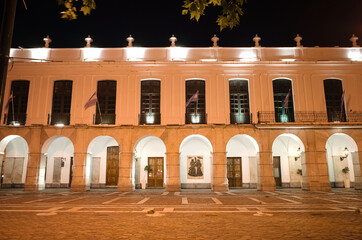 Fototapeta premium Cordoba, Argentina - January, 2020: Part of city hall of Cordoba building called Cabildo de Cordoba at night with lighting and Argentinian flags waving in wind. Old historic building with archway