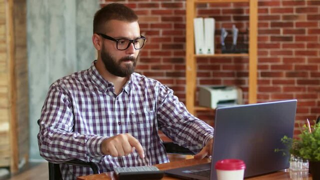 Male student with disabilities is sitting on wheelchair at table with laptop, using it, counting on calculator. Disabled hipster is trained remotely. Concept of remote education