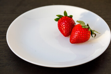 close up and side view of two large fresh red sweet strawberries on white ceramic plate on wooden black background, horizontal, copy space.