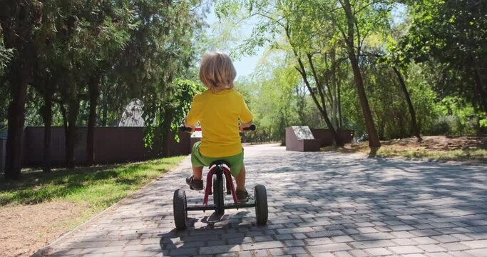 A child in a yellow T-shirt rides a tricycle along the road in the park to the fountain. The blonde boy rides a bicycle in the park.