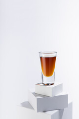 Golden scotch in shot glass in sunlight with shadows on white square podium with reflections in soft light abstract space in minimal modern style, vertical.