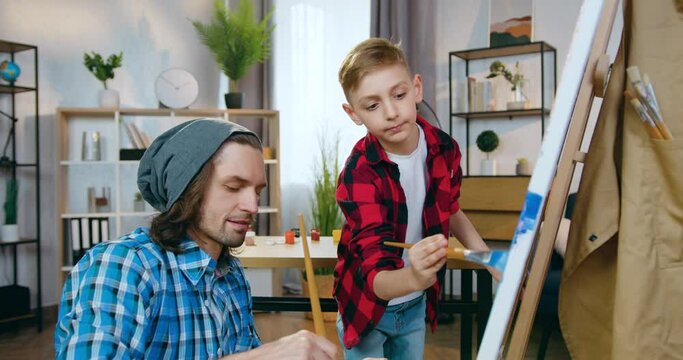 Family leisure concept where attractive positive modern dad-artist drawing picture together with his cute 10-aged son in home workshop