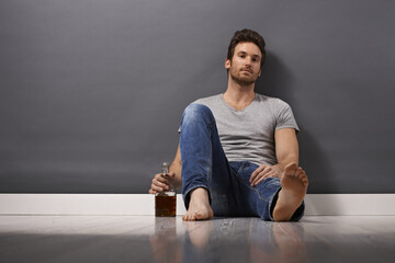Young man with bottle of whiskey sitting on floor, drinking. - obrazy, fototapety, plakaty