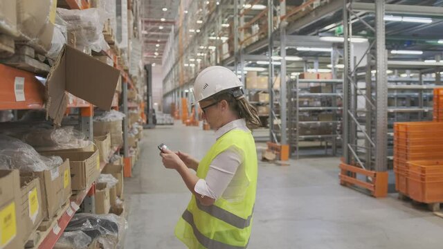 Woman manager or storekeeper using tablet for inventory control while standing near rack with cardboard boxes with goods.