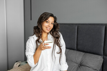 A young adult girl drinks coffee in the morning in bed. Brown-haired woman smiling.