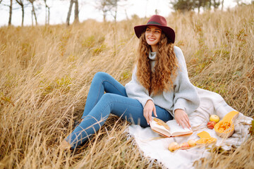 Fall picnic in nature. Young woman sitting on the blanket reading a book and smiling outdoors. People, lifestyle, relaxation and vacations concept.