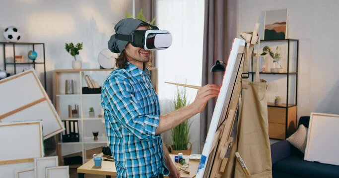 Attractive smiling talented bearded painter in virtual reality headset drawing picture on canvas in home studio