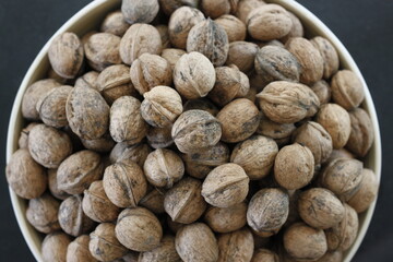 Closeup of a bunch of self-harvested homegrown whole unpeeled healthy walnuts with shell stacked in a white bowl used for decoration and food snack on a black granite countertop background in top view