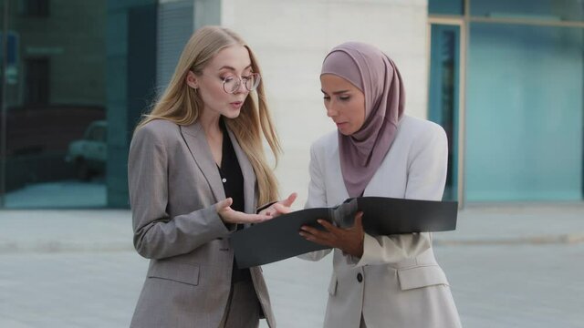 Young Indian woman in hijab outraged by report mistakes, angry scolds young intern European millennial girl with glasses. Caucasian female office worker guilty apologize for incorrectly document
