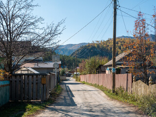 Fototapeta na wymiar Chemal, Altai Republic, Russia - 10 October 2020: Village road along wooden houses and and fence. Autumn view against the backdrop of mountains and blue sky.